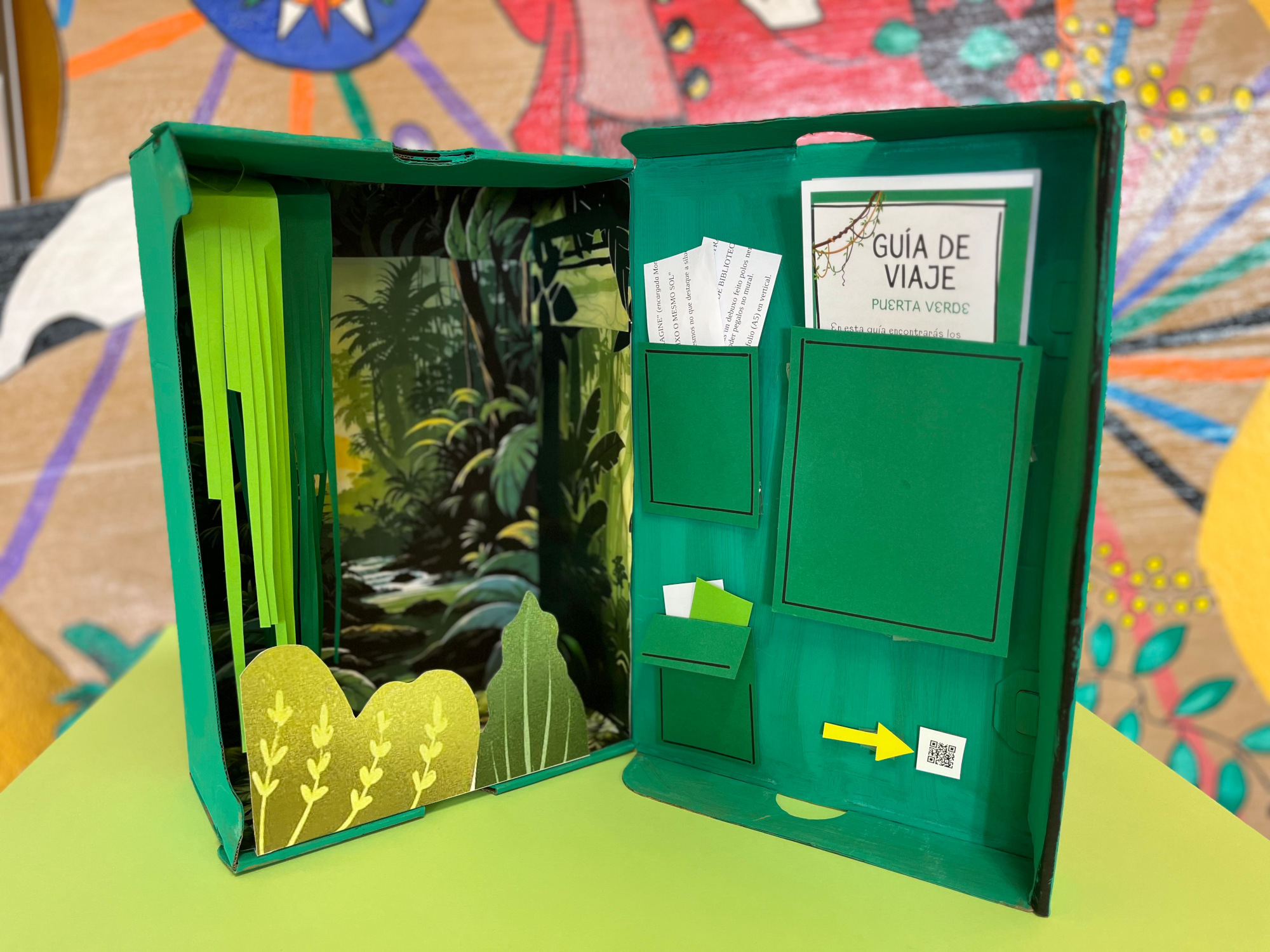 Caja de color verde con la tapa abierta. En su interior se ve una selva. En la tapa diferentes bolsillos con documentos 