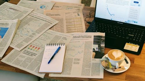 Mesa de escritorio con hojas de periódico llenas de gráficas, mesa de ordenador, un café y libreta de notas.