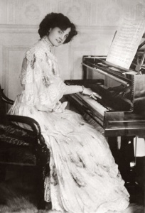 Imaxe antiga dunha muller cun vestido claro sentada tocando o piano e mirando á cámara.