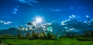Fotografía dun día de nubes e claros nunha paisaxe de campos verdes con montañas ao fondo.