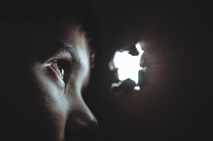 Fotografía de un primer plano de la cara en sombra de un niño observando con curiosidad por un agujero en una pared. 