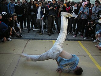 Foto dun b-boy bailando breakdance no chan frente a unha multitude