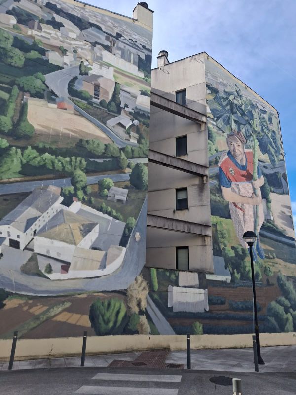 Mural de una anciana subiendo por el tronco de una berza con huertas de fondo en la fachada de un bloque de viviendas