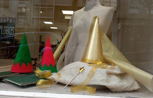 Escaparate de una sombrerería. Hay un maniquí, un sombrero de hada y dos sombreros de elfo. Los sombreros tienen forma cónica.