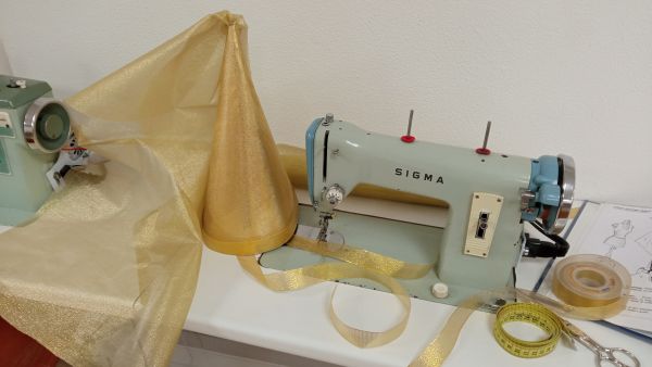 Sombrero de hada en color dorado en una mesa de trabajo de una sombrerería. Se ve una maquina de coser, tela, cinta dorada, tijeras, cinta métrica...