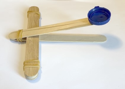 catapulta_paso3 (cREAgal) Tercer paso para construir la catapulta. Palos de madera colocados en T y con gomas atadas y tapón colocado en la parte superior.