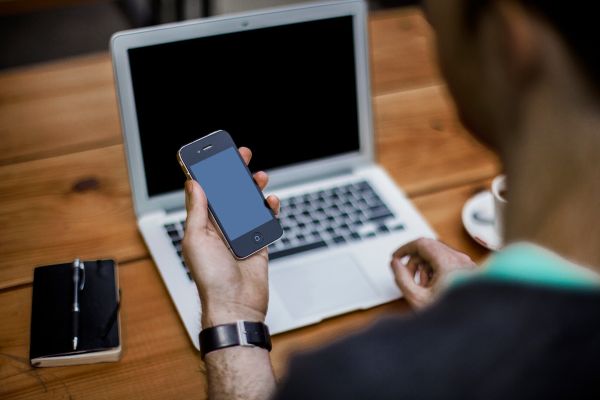 Man with smartphone and laptop (Unsplash). Una persona mira su smartphone sentada delante de su ordenador portátil.